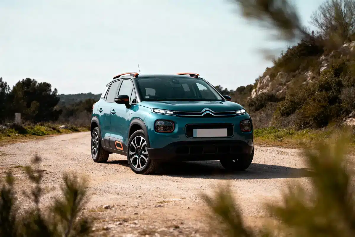 Voiture Citroën bleu sur chemin de terre, entourée de végétation et collines en arrière-plan, ciel clair.