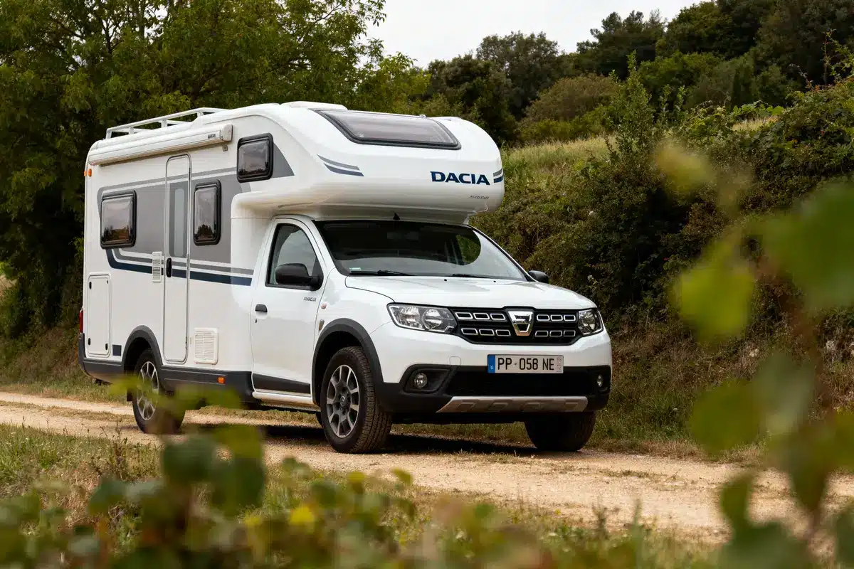Camping-car blanc Dacia Stationné sur chemin terre bordé d’arbustes, nature verdoyante en arrière-plan.