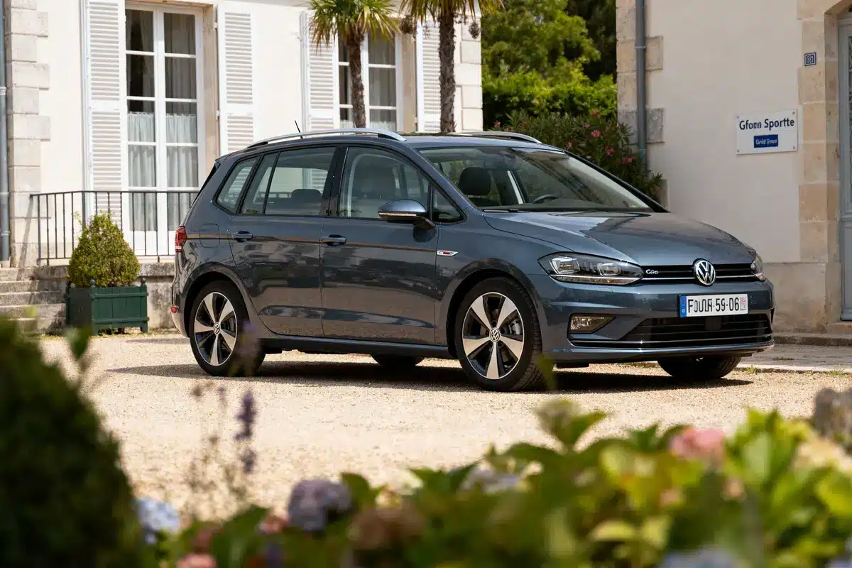 Voiture Volkswagen grise stationnée devant maison blanche avec volets, plaques françaises, jardin fleuri au premier plan.