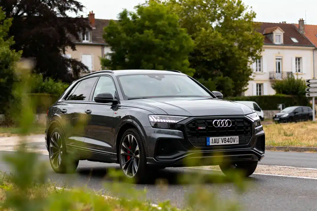 SUV Audi gris sur route, maisons en arrière-plan, verdure au premier plan, journée nuageuse.