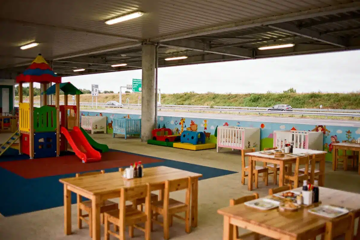Aire de repos couverte, aire de jeux colorée, tables en bois, route visible à l'arrière-plan.