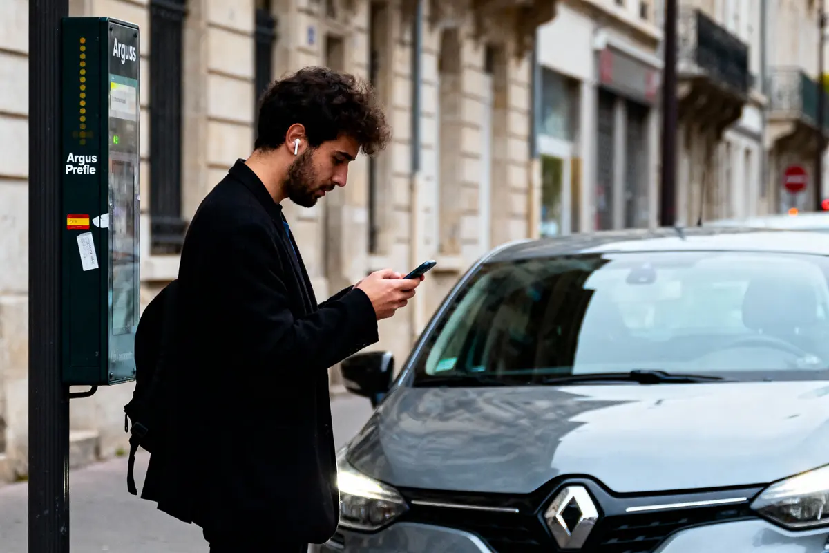 Homme avec écouteurs, utilisant un téléphone à côté d'une voiture Renault grise dans une rue urbaine.