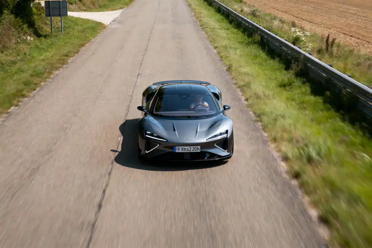 Voiture sport grise sur route campagne, devant champs et barrières métalliques, vue aérienne.