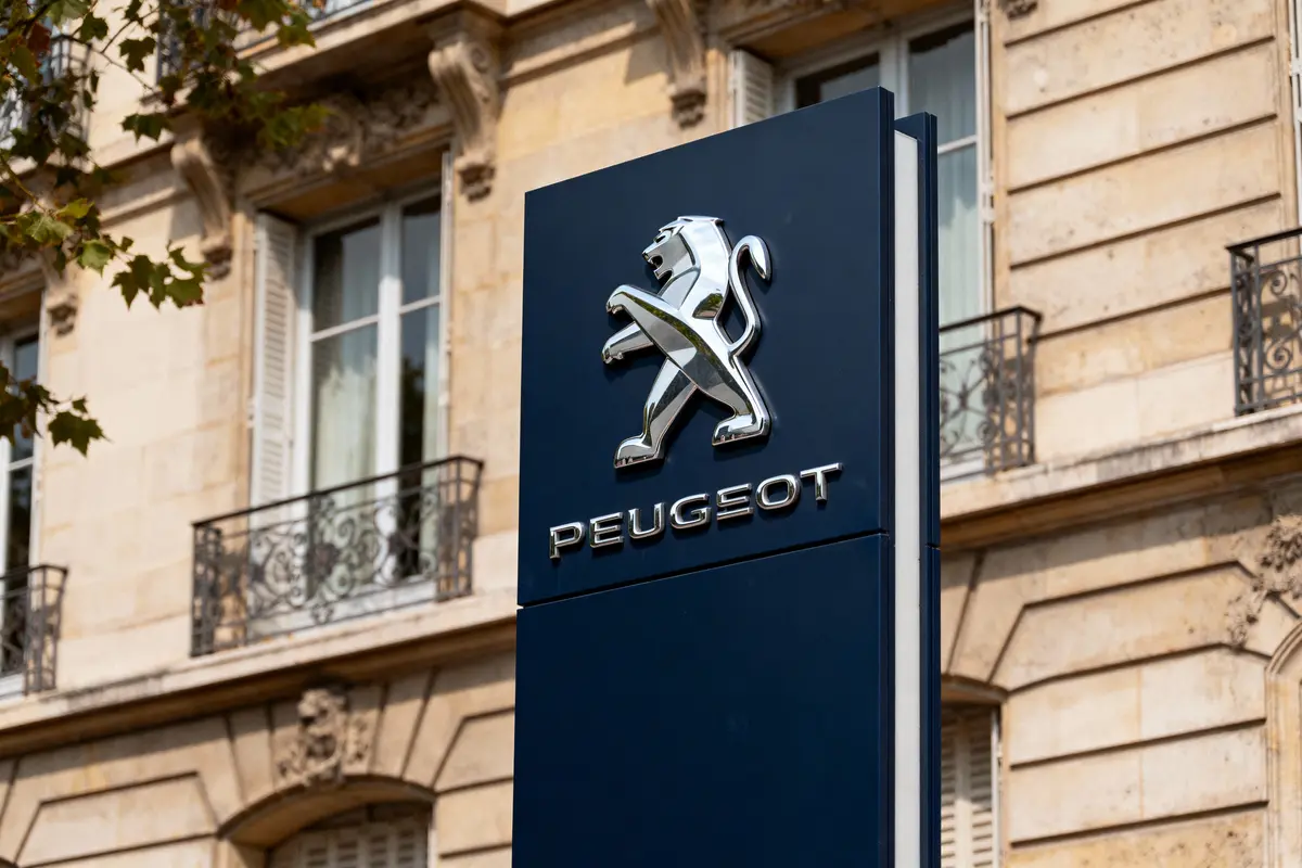 Panneau Peugeot bleu avec lion et bâtiment en arrière-plan, fenêtres et balcons ornés visibles.