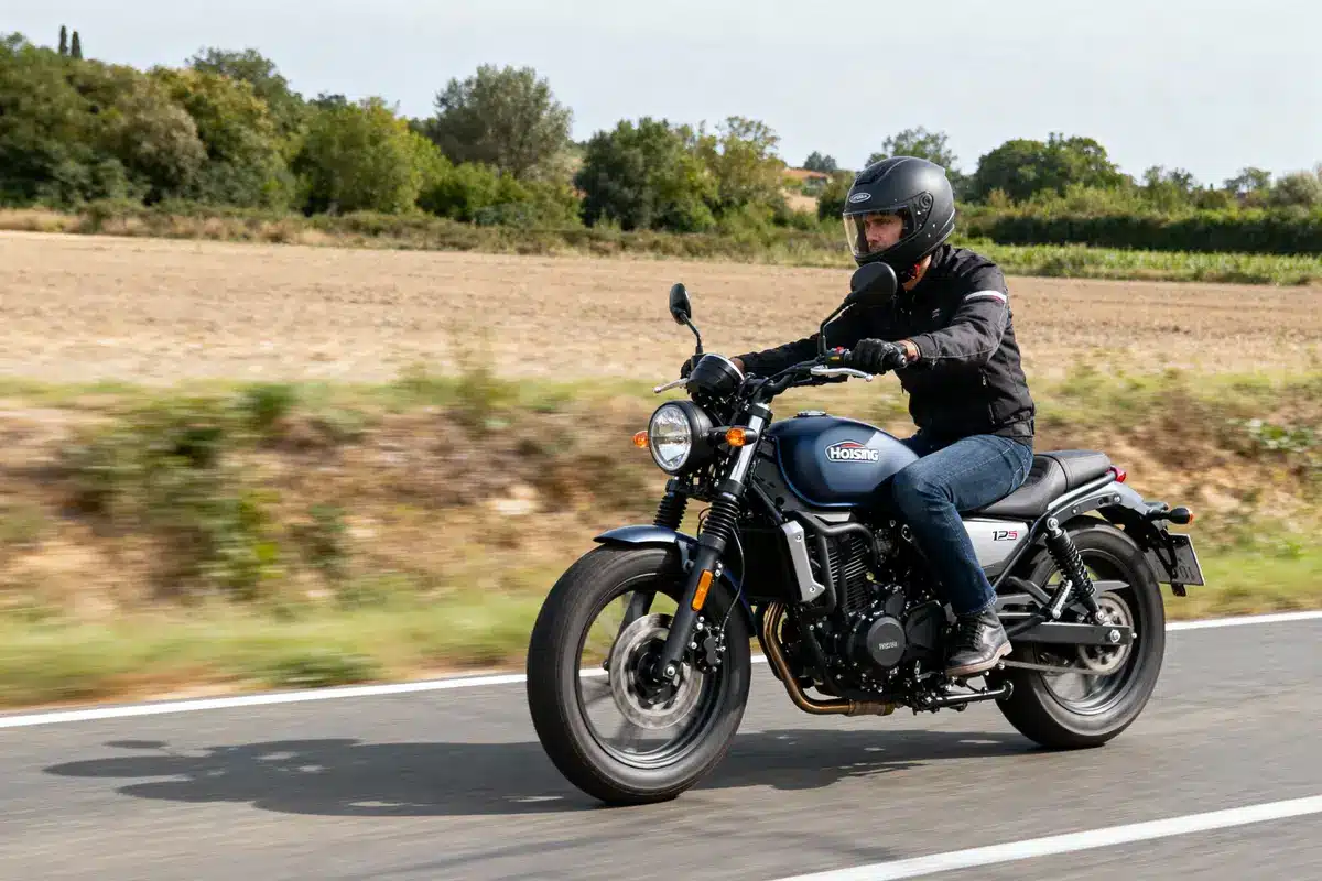Un motocycliste roule sur une route de campagne, portant un casque et des vêtements de protection noirs.