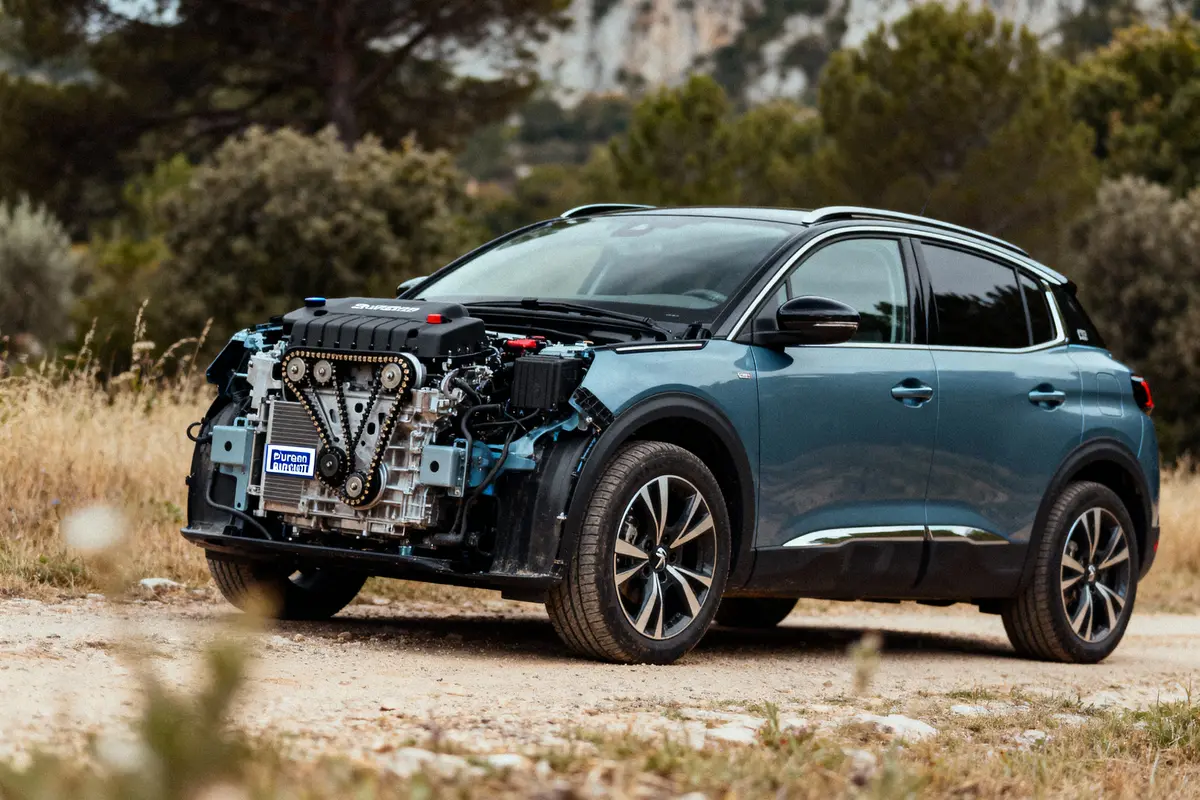 SUV bleu avec moteur à l'air libre, garé sur chemin bordé d'herbes, arbres en arrière-plan.