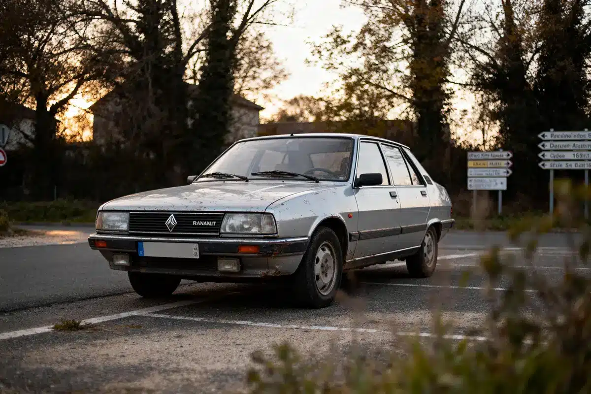 Vieille voiture grise-bleue sur une route, entourée d'arbres et panneaux, au crépuscule.