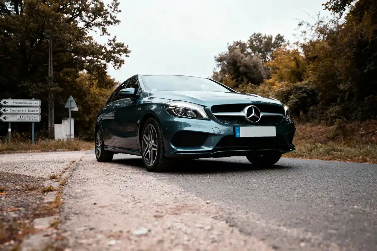 Voiture Mercedes bleu sur route de campagne, entourée d'arbres, panneaux de signalisation visible en arrière-plan.