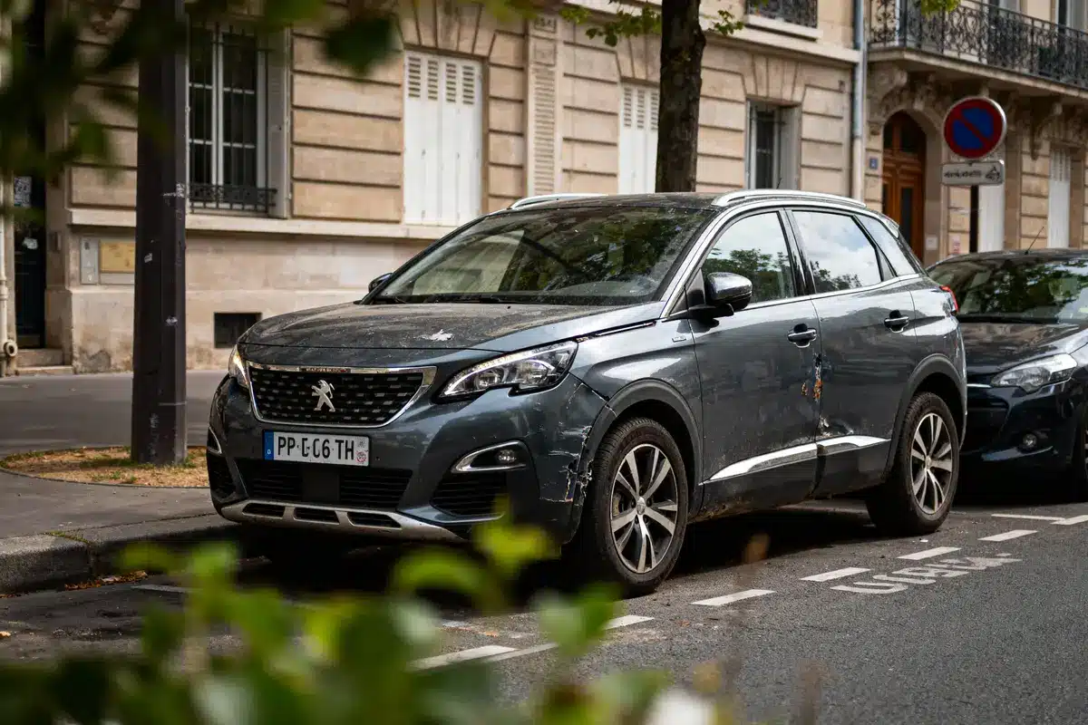 SUV gris, légèrement endommagé, stationné dans une rue urbaine avec bâtiments en arrière-plan. Feuillage flou au premier plan.