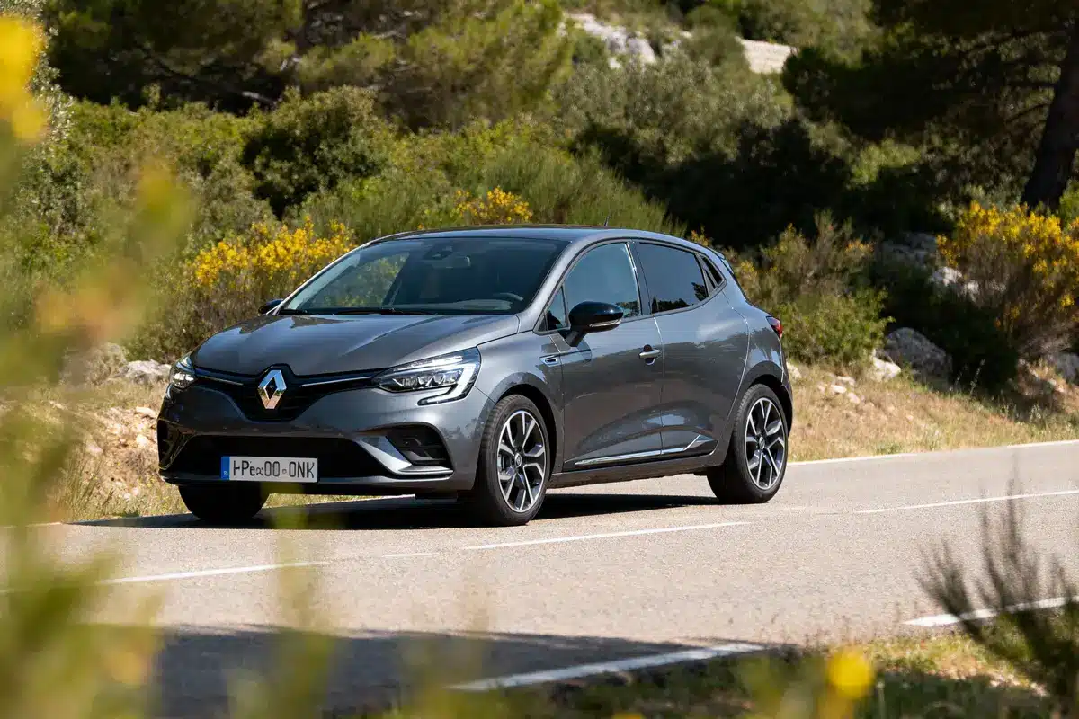 Voiture grise sur route ensoleillée, entourée de végétation et de fleurs jaunes.