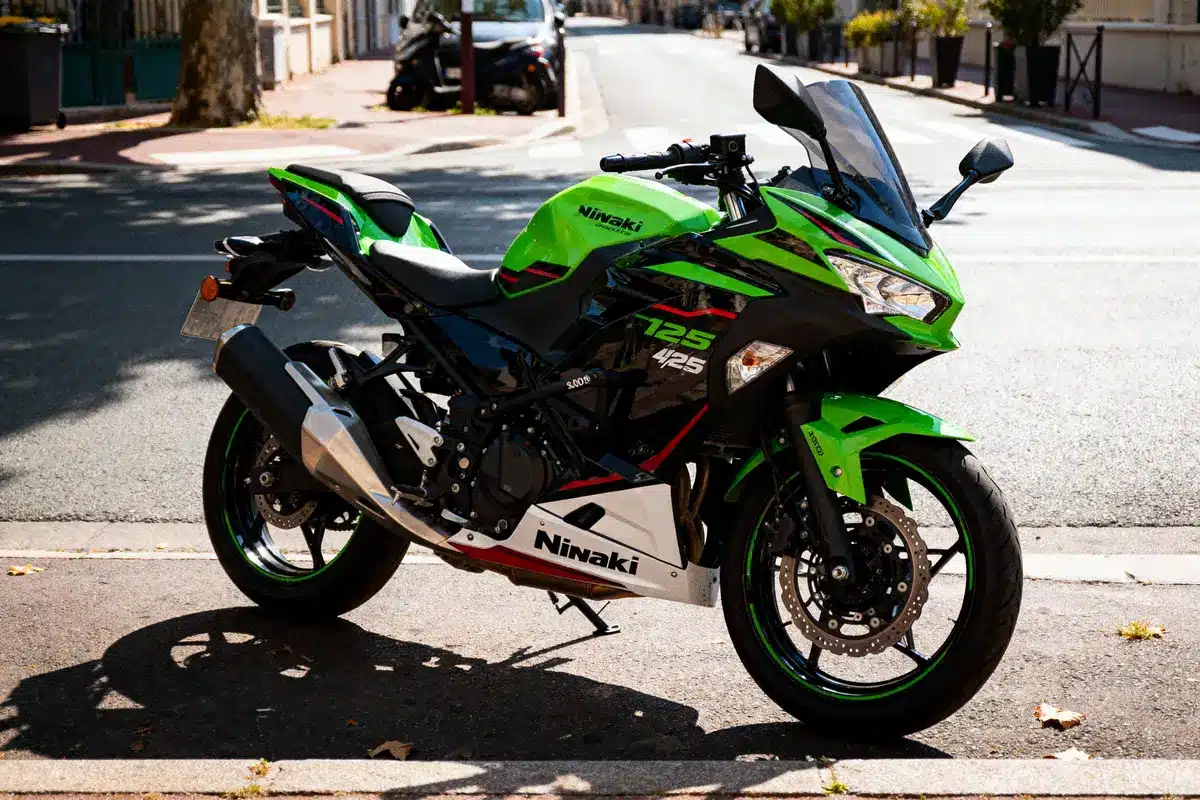Moto verte et noire garée sur trottoir ensoleillé, avec un pot d'échappement argenté et un fond urbain.
