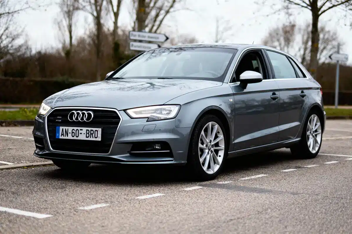 Voiture grise Audi A3 stationnée sur parking, arbres et panneaux en arrière-plan, journée nuageuse.