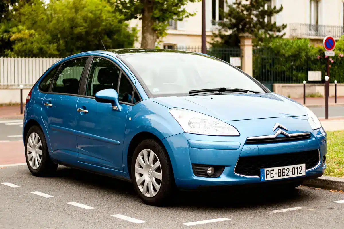 Voiture Citroën C4 bleue stationnée en bord de route, devant des arbres et bâtiments résidentiels.