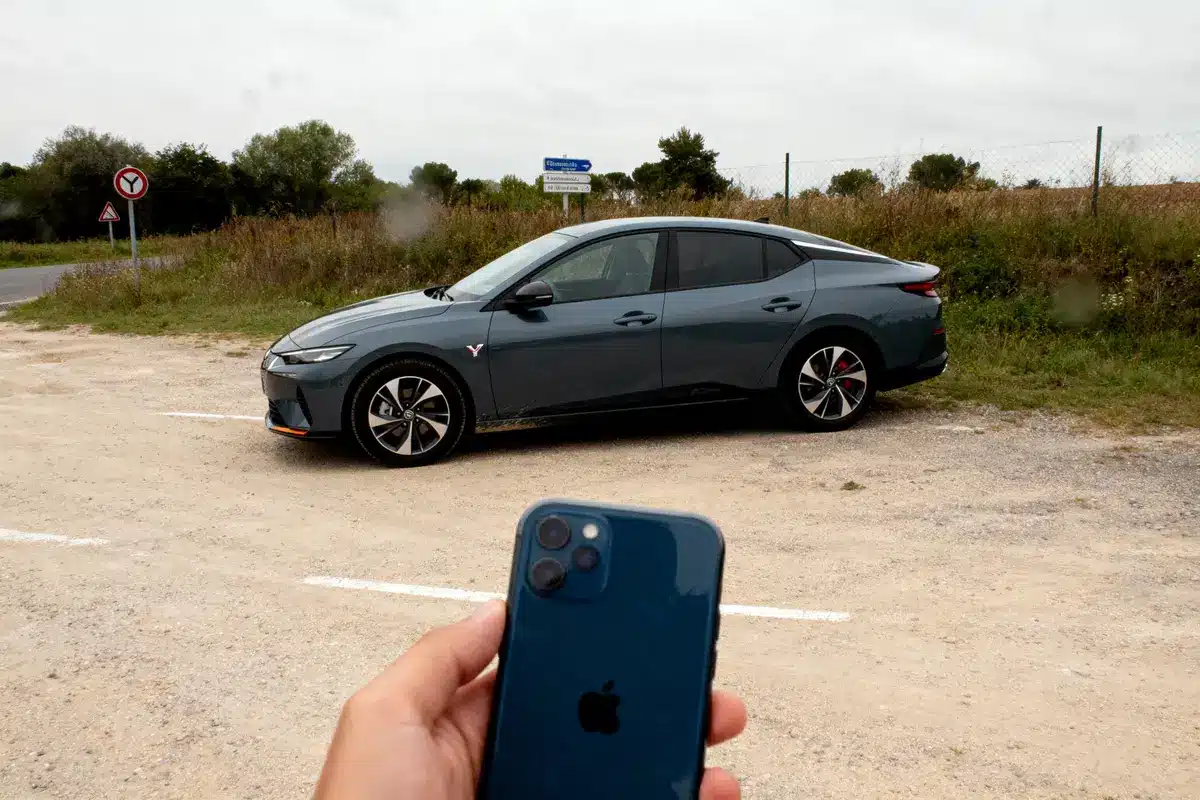 Main tenant un smartphone devant une voiture grise garée sur un chemin, panneaux routiers en arrière-plan.