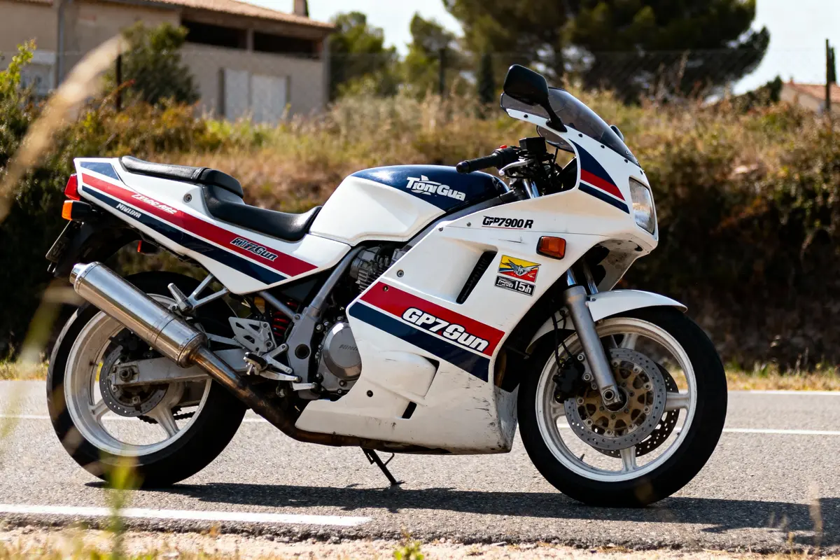 Moto blanche GPZ 900R stationnée sur une route, entourée de végétation et bâtiment en arrière-plan.