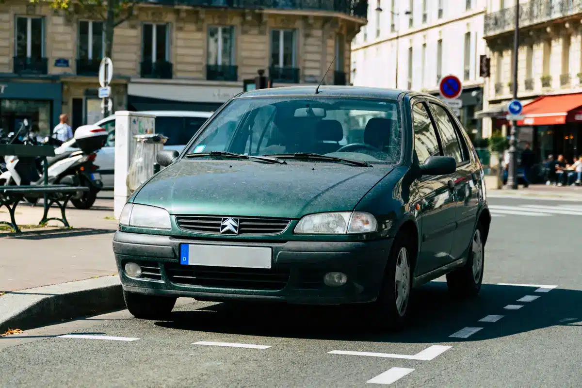 Voiture Citroën verte garée en ville, sous le soleil, près d'un bâtiment haussmannien et de scooters.