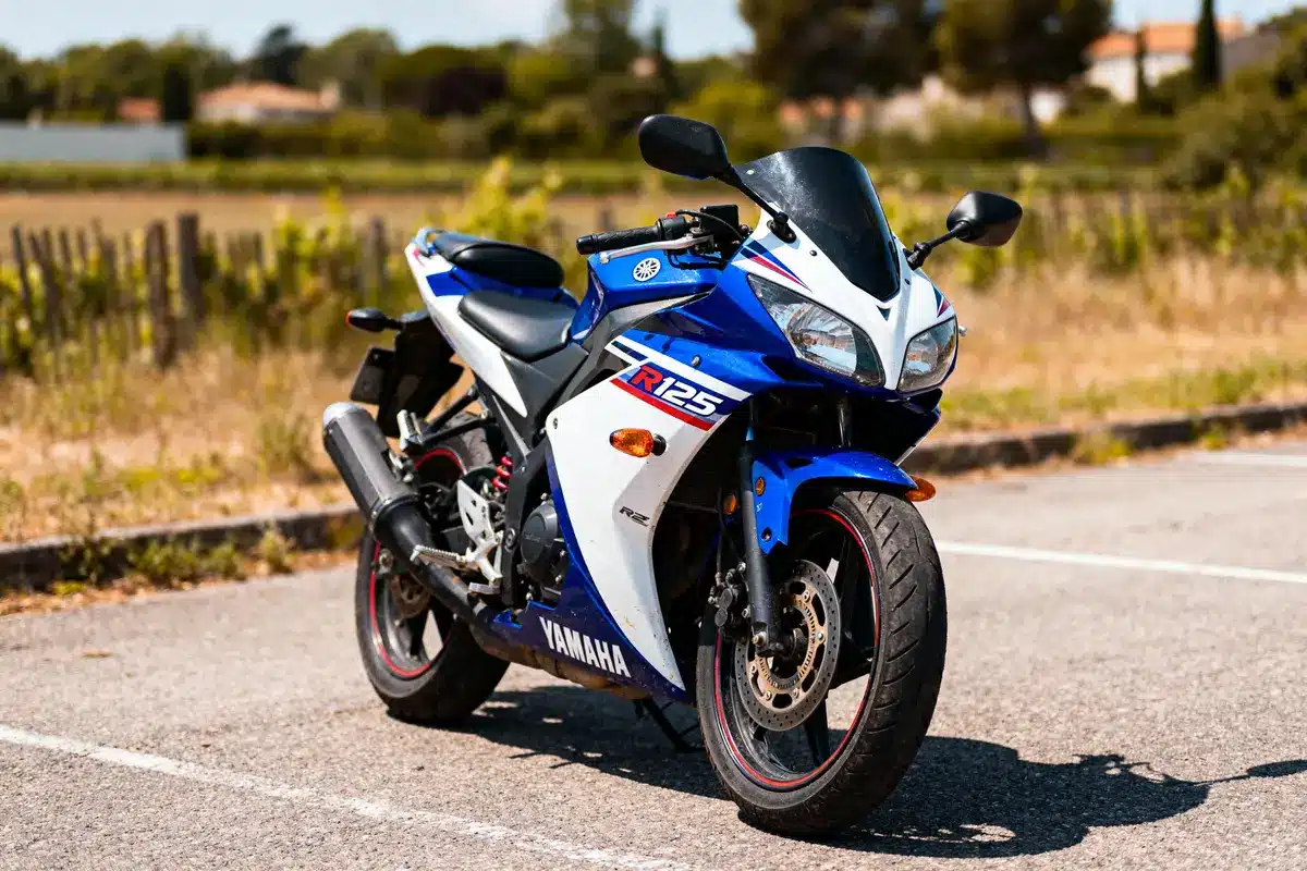 Moto sportive bleue et blanche Yamaha R125 stationnée sur route, arrière-plan champ et arbres flous.