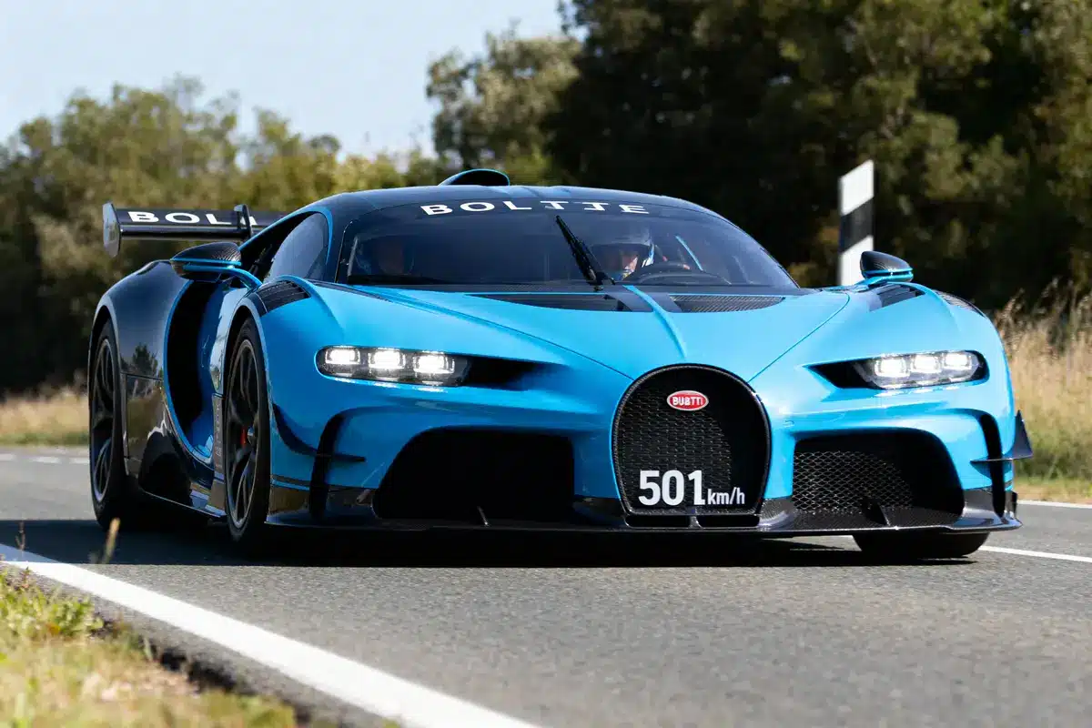 Voiture de sport bleue portant l'inscription "501 km/h" sur une route bordée d'arbres.