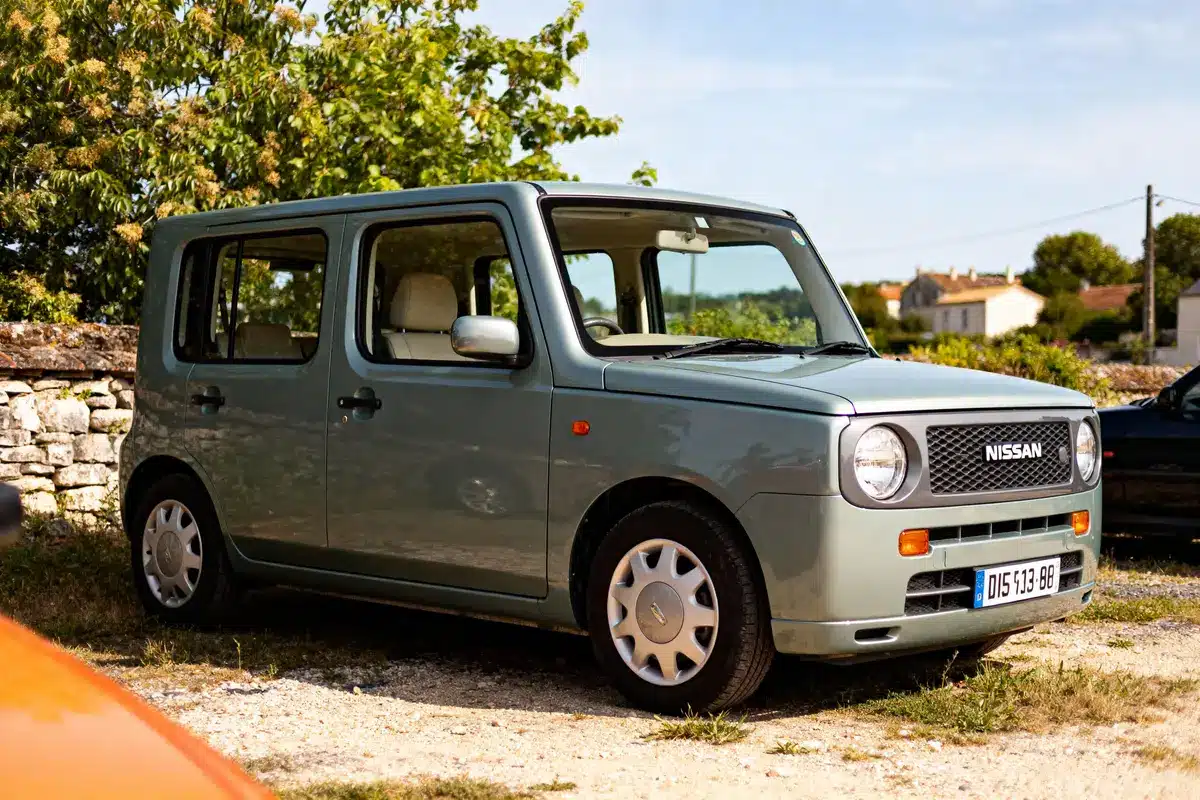 Voiture Nissan grise stationnée devant mur de pierre, avec des arbres en arrière-plan par temps ensoleillé.