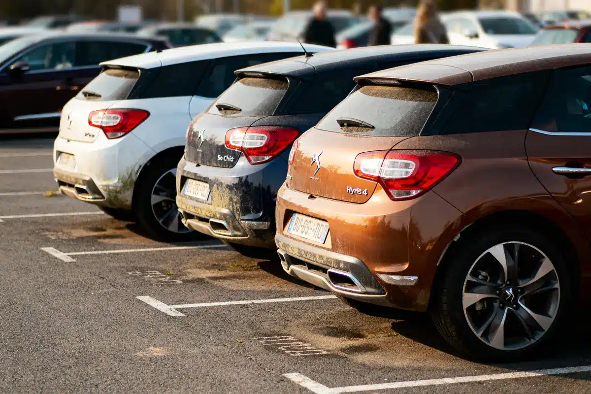 Trois voitures de couleur blanche, grise, marron garées en ligne dans un parking extérieur.