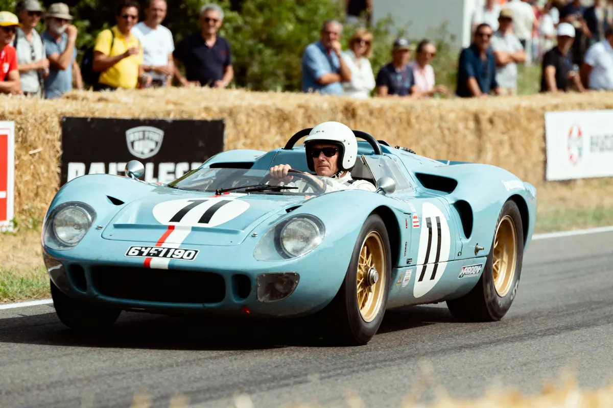 Voiture de course bleue sur circuit, pilote portant casque, spectateurs en arrière-plan. Gigantesques bottes de foin décoratives.