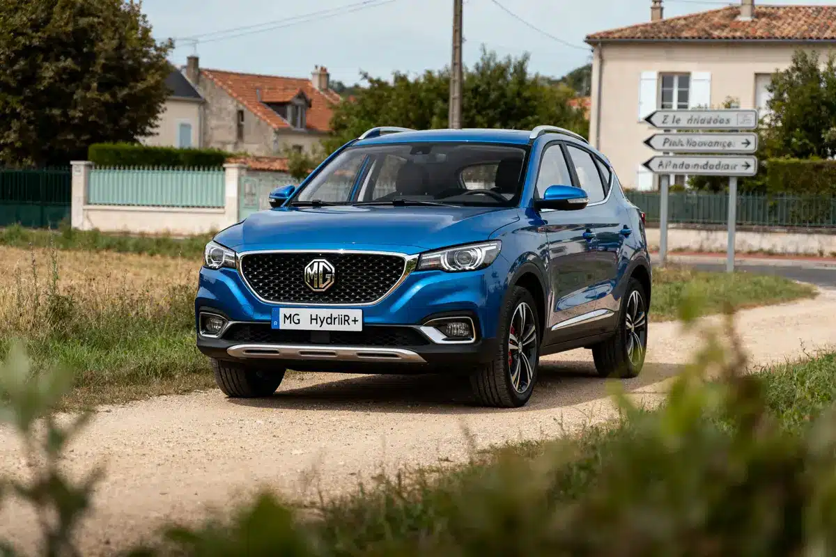 Voiture MG bleue sur chemin rural, maisons et panneaux routiers en arrière-plan.