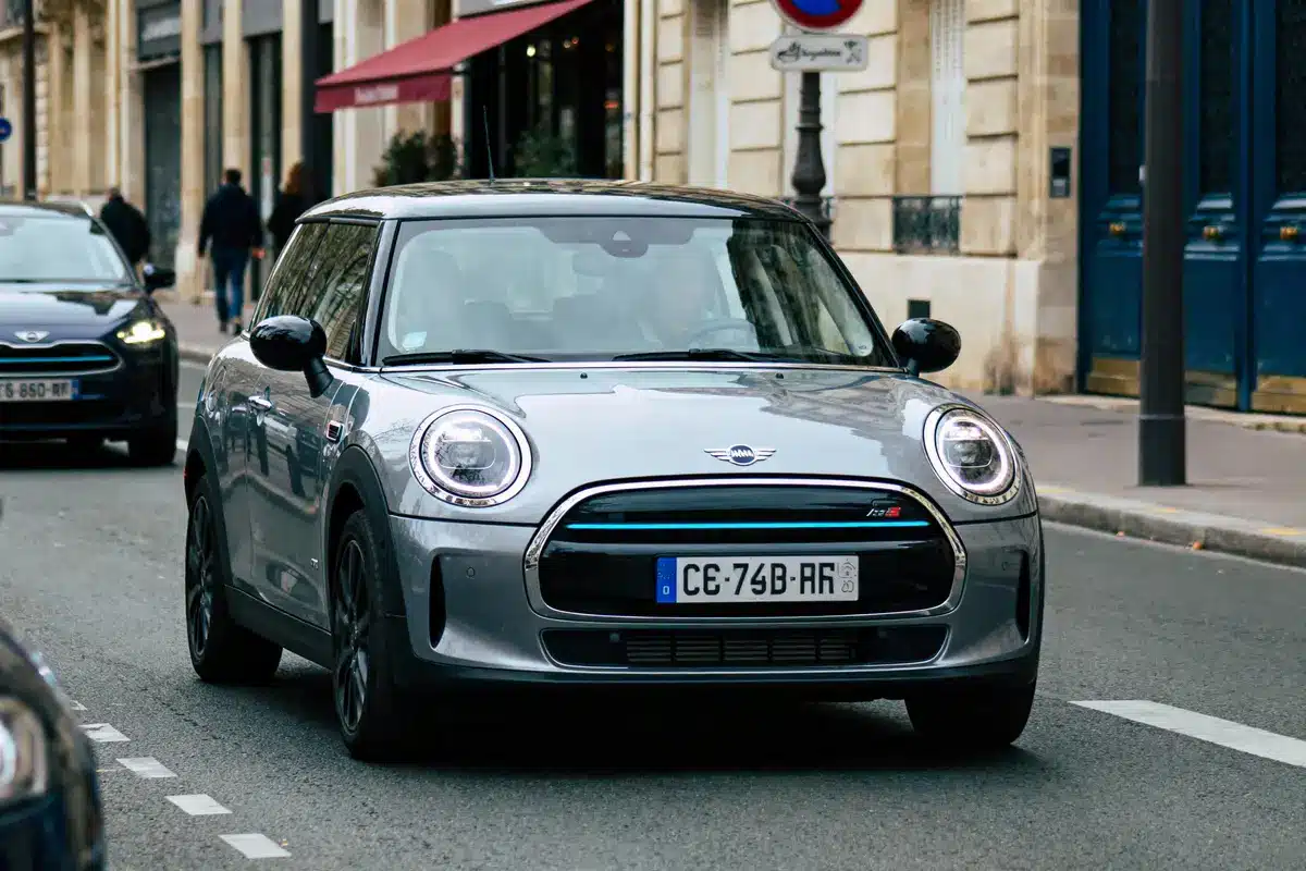 Voiture Mini grise roulant en ville, rue bordée de bâtiments anciens et piétons, jour ensoleillé.