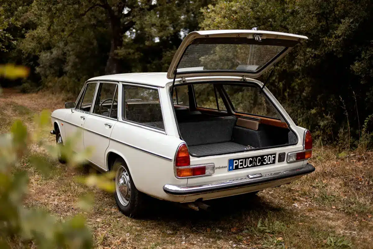 Voiture break vintage blanche sur un chemin forestier, coffre ouvert, nature environnante verdoyante.