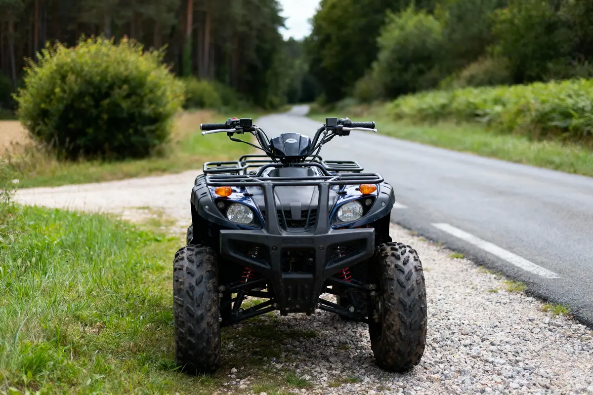 Quad noir garé sur un chemin de gravier, bordé de verdure, forêt en arrière-plan.