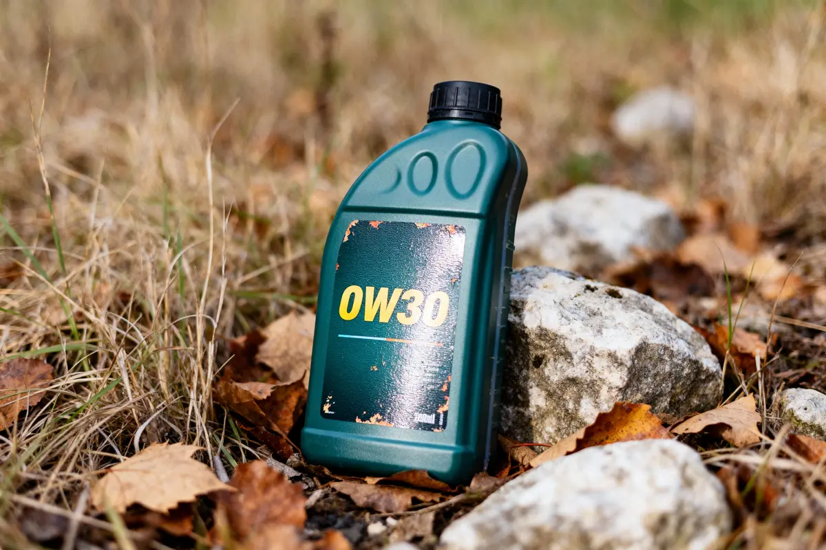 Bidon d'huile moteur 0W30 vert, posé sur des pierres, herbes sèches et feuilles mortes.