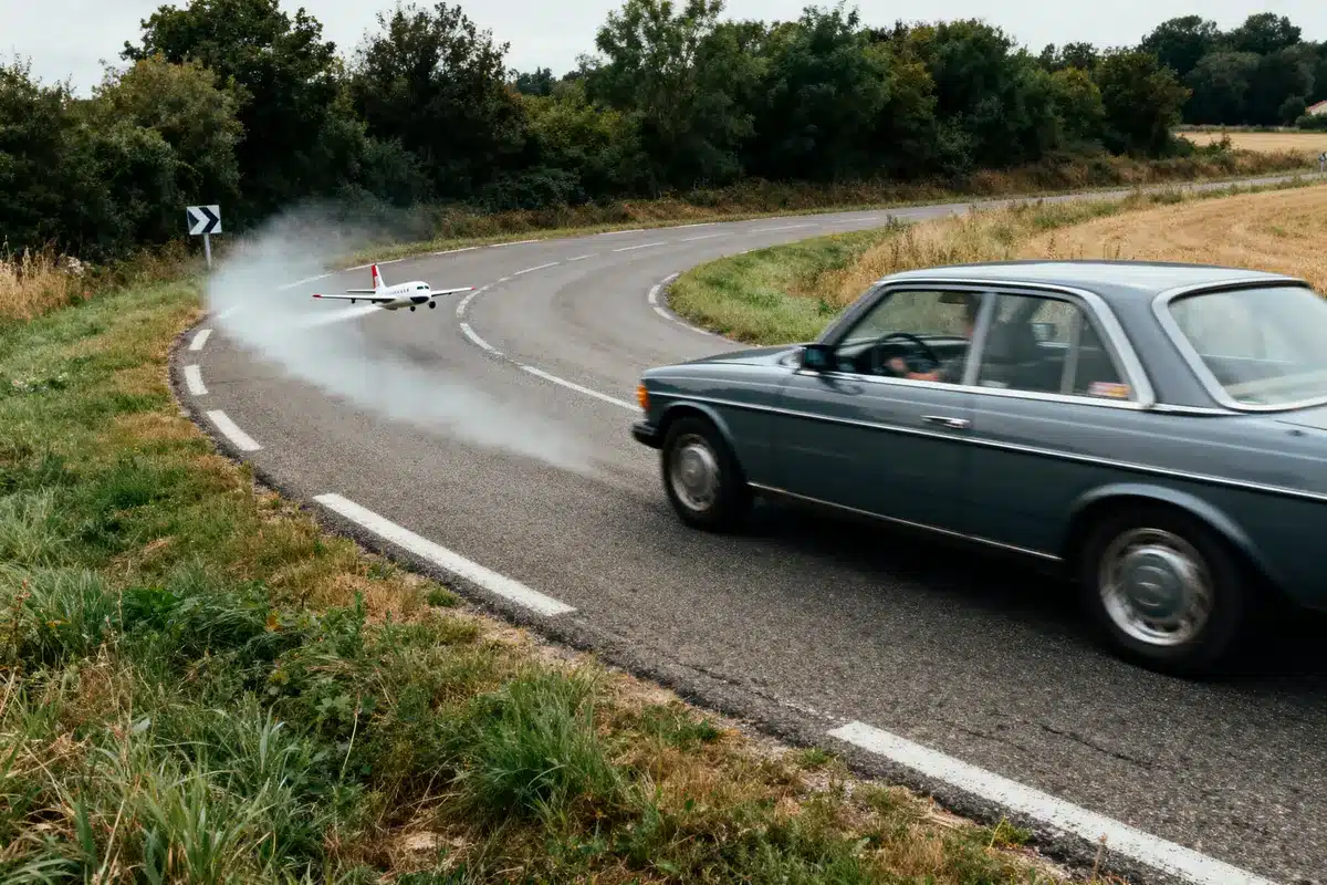 Voiture rétro sur route sinueuse avec avion miniature volant bas, laissant une traînée de fumée.