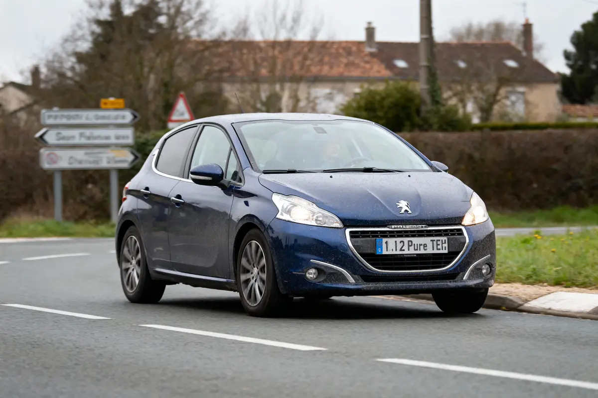 Voiture Peugeot bleue circulant sur route avec panneaux directionnels en arrière-plan.