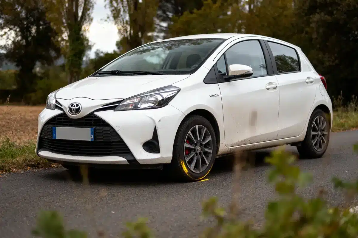 pexels-michal-pech-213601-757318 (1) Voiture blanche compacte Toyota Yaris, garée sur une route campagnarde bordée d'arbres et de végétation.