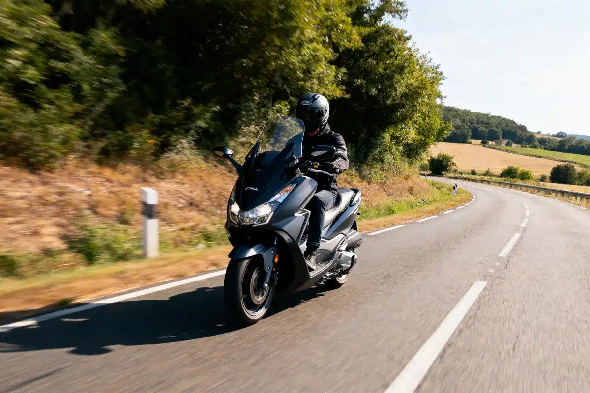 Motard en scooter noir roulant sur une route de campagne sinueuse, journée ensoleillée, forêt en arrière-plan.