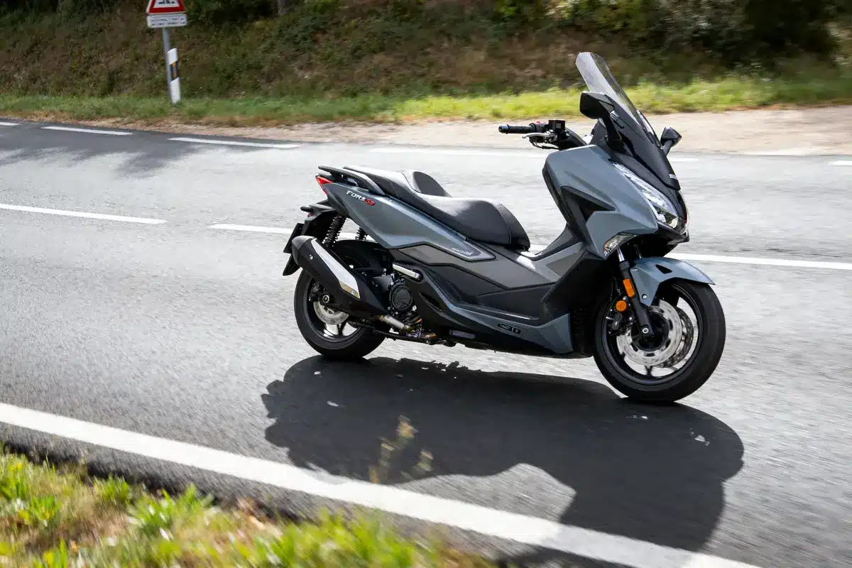 pexels-michal-pech-213601-757318 (1) Scooter gris roulant sur route, paysage verdoyant en arrière-plan, panneau de signalisation visible.