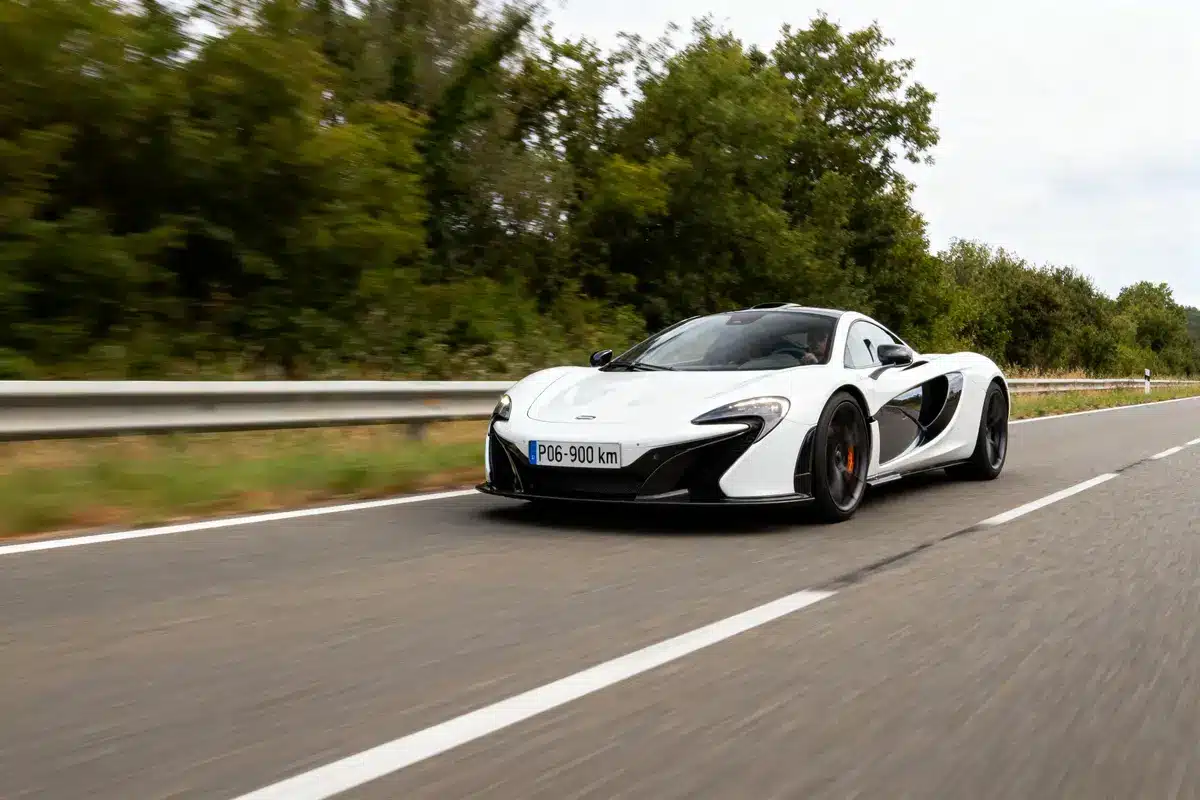 Voiture de sport blanche roulant sur route, entourée de verdure, ciel nuageux. Plaque: "PO6-900 km".