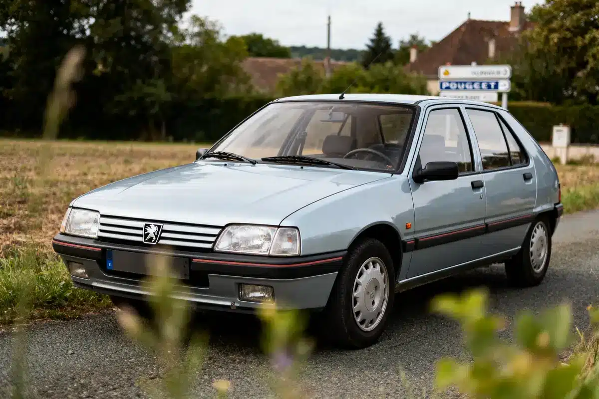 Voiture Peugeot bleue garée sur une route de campagne, panneau de signalisation visible à l'arrière-plan, nature environnante.
