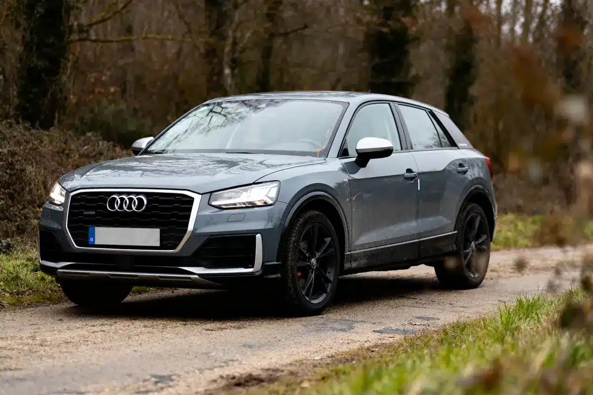 SUV gris Audi garé sur chemin rural, entouré d'arbres, en automne.