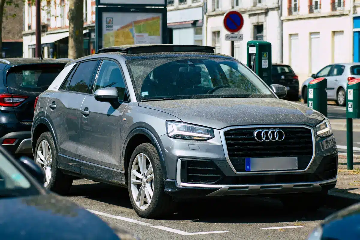SUV gris Audi garé dans la rue, légèrement couvert de poussière, à côté de bornes de recharge électriques.