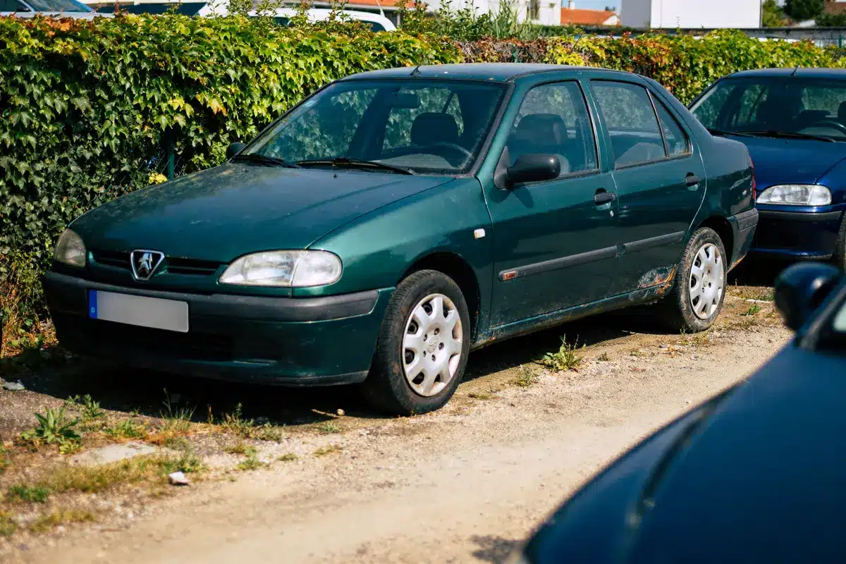 Voiture verte garée sur un sol en terre, entourée de végétation et d'autres voitures.