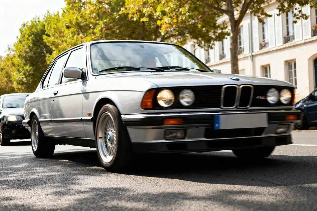BMW e28 automatic grise roulant dans une rue bordée d'arbres et d'immeubles élégants