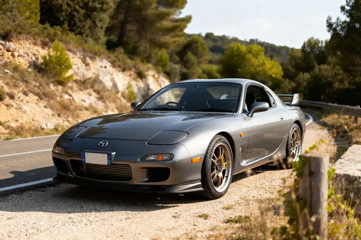 Mazda RX7 coupé gris garé sur route sinueuse bordée d'arbres et collines